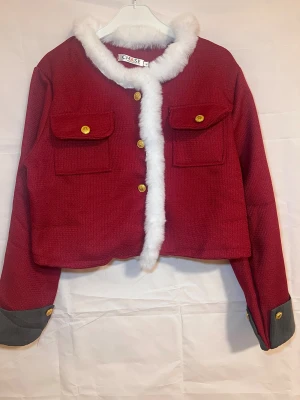 Vinröd jacka med fuskpäls  - Säljer en röd pälsjacka med vit fuskpäls runt krage och knappslå. Jackan har två stora fickor med guldknappar framtill och gråa muddar med gulddetaljer vid ärmslut. Croppad passform och långärmad, perfekt för att sticka ut.