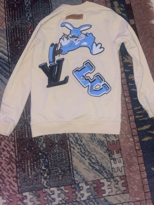 Louis Vuitton beige sweatshirt med tryck - Säljer en beige sweatshirt från Louis Vuitton i 100% bomull. Tröjan har lång ärm och coolt tryck med blå kanin och LV-logga på ryggen samt blå LV-märke på bröstet. Perfekt för dig som gillar streetwear med lyxig touch och snygga detaljer.