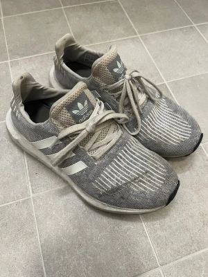 Grå adidas sneakers strl 44 - Säljer ett par grå adidas sneakers med vita ränder och mesh-ovandel. Skorna har snörning och en platt sula, samt adidas logga på tungan. Mönstret är diskret randigt och materialet är syntet och mesh. Perfekt för dig som gillar sportig stil.