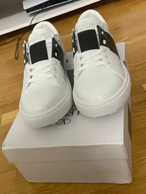 Valentino Garavani vita sneakers - Snygga vita sneakers från Valentino Garavani med svart detalj och nitar på sidan. Skorna har rund tå, snörning och en chunky platt sula. Tillverkade i slätt skinn med guldfärgad logga på insidan. Perfekta för dig som vill ha en clean men lyxig look.