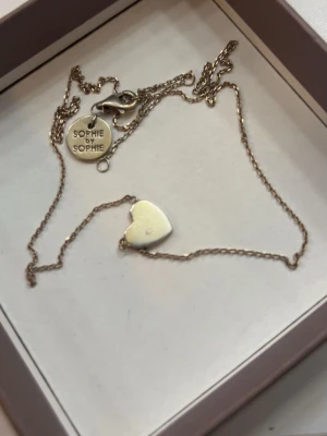 Guldhalsband med hjärta Sophie by Sophie - Säljer ett tunt och elegant guldhalsband från Sophie by Sophie med ett stilrent hjärthänge. Kedjan är finlänkad och har en liten rund bricka med varumärket ingraverat vid låset. Perfekt för dig som gillar minimalistiska smycken.