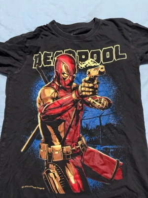 Svart Deadpool t-shirt och joker med tryck (2st) - Cool svart t-shirt med stort Deadpool-tryck i rött, gult och blått framtill. Klassisk passform med rund hals och korta ärmar. Perfekt för dig som älskar Marvel och vill sticka ut med en färgstark och ikonisk design.