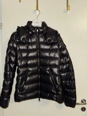 moncler dam bady jacka  - Säljer en svart pufferjacka från Moncler med glansig finish och huva. Jackan har dragkedja framtill, två sidofickor med dragkedja och Moncler-logga på ärmen. Perfekt för kalla dagar och har en riktigt snygg streetstil.  storlek 3 (medium)