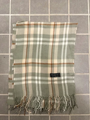 Rutig ullhalsduk från Burberrys - Klassisk rutig halsduk från Burberrys i mjuk lammull. Färgkombinationen går i beige, grått, vitt och brunt med fransar i ändarna. Perfekt accessoar för att lyfta din outfit med en tidlös brittisk känsla.