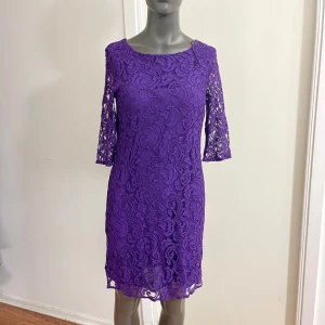 Lila spetsklänning med trekvartsärm - Superfin lila klänning i spets med trekvartsärmar och rund halsringning. Klänningen är kort och har ett vackert blommigt spetsmönster som ger en elegant och trendig look. Perfekt för dig som vill sticka ut med färg och stil.