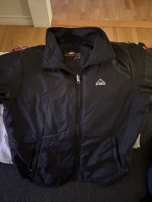 Svart pufferjacka från McKinley - Svart pufferjacka från McKinley med hög krage och hel dragkedja framtill. Jackan har två sidofickor med dragkedja och en stilren design utan mönster. Perfekt för kyliga dagar och enkel att matcha med din vardagsstil.