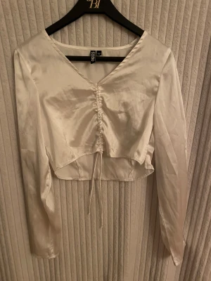 Vit croppad blus från Pieces - Säljer en vit croppad blus från Pieces i glansigt satinmaterial. Toppen har v-ringning, långa ärmar och snygg snörning framtill som ger en cool rynkad effekt. Använt en gång och är i mycket bra skick. 