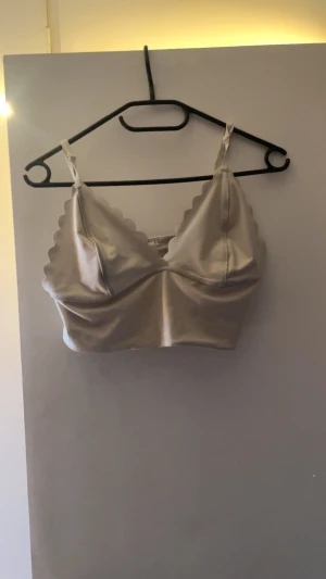 Champagnefärgad satin bandeau topp - Champagnefärgad bandeau topp i satin med smala axelband och vågiga kanter. Snygg och stilren design som ger en lyxig känsla. Perfekt att styla med höga jeans eller kjol för en trendig look.