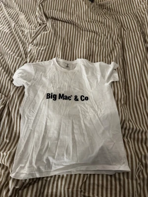 Vit t-shirt Big Mac - Säljer en vit t-shirt med svart text 'Big Mac & Co' på framsidan och ett coolt tryck med '85 kr' i rött och vitt på ryggen, från Mcdonalds. T-shirten har klassisk rund hals och korta ärmar.  Storlek S