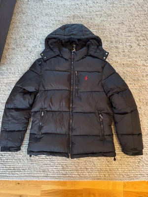 Svart pufferjacka från Polo Ralph Lauren - Svart pufferjacka från Polo Ralph Lauren med huva, röda broderade loggan på bröstet och flera dragkedjefickor. Jackan har quiltad design och är tillverkad i syntetmaterial, perfekt för kalla dagar. Stängs med dragkedja framtill och har justerbara muddar.
