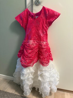 Rosa och vit prinsessklänning med volanger - Superfin långklänning med glittrig rosa överdel i blommigt spetsmönster och v-ringning. Kjolen är vit med flera lager av volanger i skimrande tyg. Klänningen har korta ärmar och dragkedja i ryggen. Perfekt för ditt barn som verkligen vill sticka ut som prinsessa i en färgstark look! Lite osäker på storleken, misstänker att den är större än 116.