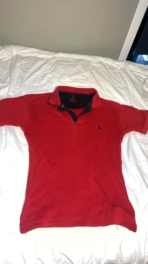 Röd pikétröja från Polo Ralph Lauren - Snygg röd pikétröja från Polo Ralph Lauren med klassisk svart logga broderad på bröstet. Tröjan har korta ärmar, svart insida på kragen och knappar framtill. Perfekt för dig som gillar stilrena och sportiga plagg med premiumkänsla.