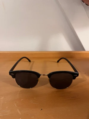 Svarta clubmaster solglasögon - clubmaster-inspirerade solglasögon med svarta bågar och guldiga detaljer. Runda, mörka glas och klassisk retrostil. 