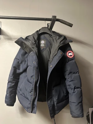 Mörkblå pufferjacka från Canada Goose - Säljer en mörkblå pufferjacka från Canada Goose med huva och klassisk logotyp på armen . Jackan har dragkedja framtill, snörning vid huvan och två stora fickor. 