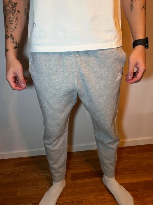 Grå Adidas mjukisbyxor med vita ränder - Snygga grå mjukisbyxor från Adidas med klassiska vita ränder längs sidorna och logga på vänster lår. Byxorna har normal passform, elastisk midja och är gjorda i mjukt bomullsmaterial. Perfekta för chill eller träning. Saknas snöre