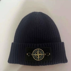 Svart ribbad mössa från Stone Island - Svart ribbstickad mössa från Stone Island med klassisk logopatch framtill. Tillverkad i mjuk ull och har en bred uppvikt kant. Perfekt för dig som vill ha en clean och stilren look med street-vibe.