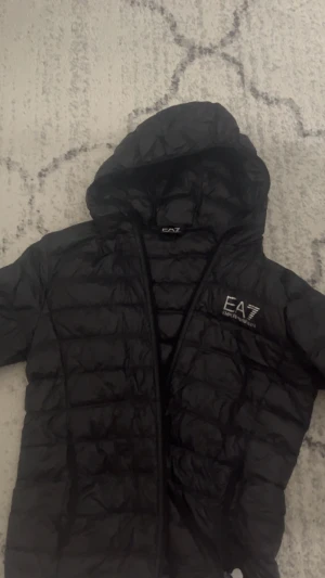 Svart pufferjacka från EA7 Emporio Armani - Svart pufferjacka från EA7 Emporio Armani med huva och dragkedja. Jackan har quiltad design och logga på bröstet. Säljer för att jag gillar inte den stilen längre! Inga defekter eller något liknande 