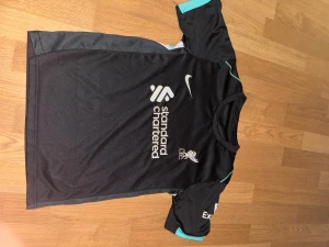 Liverpool FC svart Nike fotbollströja (cirka XS) - Säljer en svart Liverpool FC fotbollströja från Nike med vit logga och tryck från Standard Chartered. Tröjan har korta ärmar med turkosa detaljer och är tillverkad i lätt, ventilerande polyester. Perfekt för dig som vill representera LFC på planen eller läktaren.
