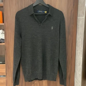 Mörkgrå pikétröja från Polo Ralph Lauren - Snygg mörkgrå långärmad pikétröja från Polo Ralph Lauren med klassisk krage och två knappar vid halsen. Tröjan har diskret logobroderi på bröstet och är tillverkad i mjukt material som känns skönt mot huden.