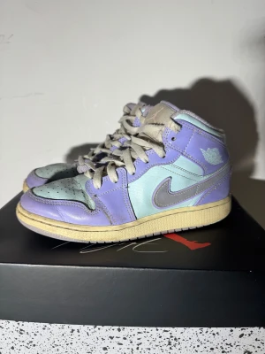 Nike Air Jordan 1 aqua 37.5 - Nike Air Jordan 1 Mid i snygga pastellfärger med ljuslila och mintgrön skinnpanel, grå swoosh och beige sula. Klassisk snörning och rund tå. Perfekt för dig som vill sticka ut med färg på fötterna. Ikonisk Jordan-logga på sidan.