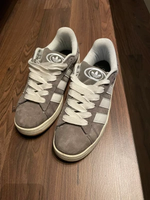 Adidas Campus grå sneakers mocka - Säljer ett par Adidas Campus sneakers i grå mocka med vita klassiska stripes och vit sula. Skorna har rund tå, platt sula och snörning. Adidas-logga på plösen och sidan. Perfekt för dig som gillar retrostil och streetwear.