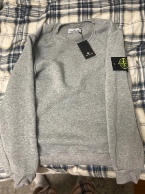 Grå Stone Island sweatshirt - Snygg grå sweatshirt från Stone Island med klassisk rund hals och ribbade muddar. Tröjan har den ikoniska Stone Island-patchen på vänster ärm med broderad logga i gult och grönt. Perfekt för dig som gillar streetwear och vill ha en clean look.