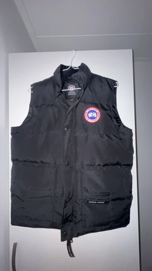 Svart dunväst från Canada Goose - Svart dunväst från Canada Goose med klassisk logga på bröstet och två stora fickor framtill. Västen har hög krage, knappar och dragkedja. Perfekt för kalla dagar och har en clean, stilren look som funkar till många outfits.