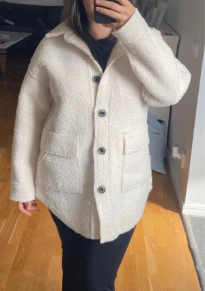 Vit teddyjacka med stora fickor - Supermjuk vit teddyjacka med stora knappar och två rejäla fickor framtill. Jackan har klassisk krage och lång ärm, perfekt för lager-på-lager. Materialet är fluffigt och ger en riktigt cozy vibe. Helt ny 
