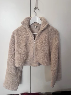 Beige croppad teddyjacka med half zip - Supermysig croppad teddyjacka i beige med hög krage och half zip framtill. Jackan har långa ärmar och är gjord i fluffigt syntetmaterial som ger en skön och chill vibe. Perfekt för kalla dagar när du vill vara både varm och snygg.