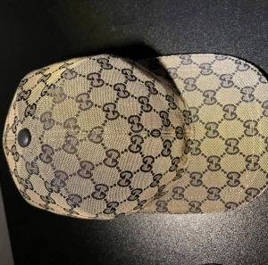 Gucci beige keps med GG-mönster - Beige-grå keps från Gucci med klassiskt GG-monogram över hela kepsen. Kepsen har en justerbar rem baktill, och den har ett rött och 2 blåa ränder på sidan. Materialet är canvas med detaljer i mörkblått tyg. Perfekt för dig som gillar lyxiga accessoarer. Box och allt som följer med medföljer såklart!!