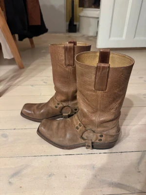 Bruna cowboy boots från Sendra - Säljer ett par bruna cowboy boots från Sendra i äkta skinn. Boots med fyrkantig tå, dekorativa metallringar och nitar runt ankeln. Klassisk westernstil med robust klack och detaljerad sömnad. Perfekta för dig som gillar statement skor med vintage vibe.