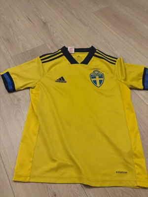 Svensk landslagströja Adidas 13-14 år - Gul fotbollströja från Adidas med Sveriges landslagsemblem på bröstet och tryck 'Filling 6' på ryggen. Tröjan har korta ärmar, blå detaljer på axlar och ärmslut samt svart V-ringad krage. Tillverkad i lätt och ventilerande Aeroready-material.