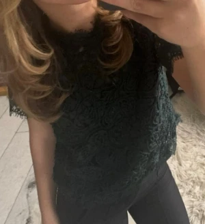 Mörkgrön spetsblus med kort ärm - Säljer en mörkgrön blus med vacker spets och korta ärmar. Blusen har en hög hals och fina detaljer längs kanterna. Perfekt för dig som gillar romantisk stil och vill ha något extra till din outfit. Står Medium, men den sitter bra på mig som är 160.