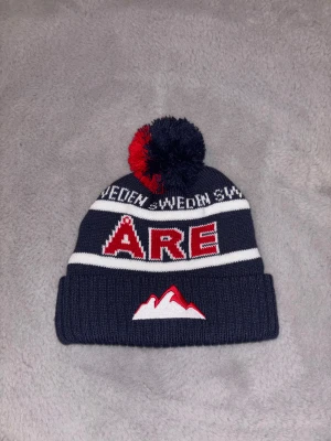 Mörkblå Åre mössa med tofs - Snygg mörkblå stickad mössa med röd och vit text 'ÅRE' framtill och broderat bergmotiv. Tofsen på toppen är i rött och mörkblått. Perfekt för kalla dagar och vinterhäng.