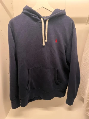 Mörkblå hoodie från Polo Ralph Lauren - Snygg mörkblå hoodie från Polo Ralph Lauren med klassisk röd broderad logga på bröstet. Tröjan har huva med vita snören, stor magficka och långa ärmar. Perfekt för chill dagar och enkel att matcha med jeans eller joggers. Skick: 10/10 har använts runt 10 gånger men säljer eftersom den är för liten på mig. Den är i storlek M men sitter mer som S. Skriv vid minsta lilla, svarar så snabbt jag kan🤗