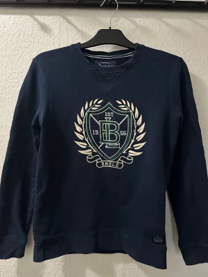 Marinblå sweatshirt från Bondelid XS - Snygg marinblå sweatshirt från Bondelid i storlek XS. Tröjan har ett stort tryck med sköld och krans på bröstet, samt ribbade muddar vid ärmslut och nederkant. Mjuk bomullskvalitet och klassisk rund hals. Perfekt för en chill och sportig look.