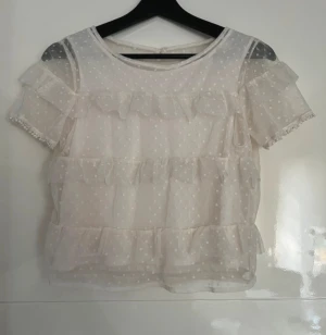 Vit prickig blus med volanger - Säljer en superfin vit blus med prickigt mönster och volanger på både fram- och baksida. Blusen har korta ärmar med små bollar längs kanten och är gjord i ett tunt, transparent material som ger en luftig känsla. Perfekt för dig som gillar romantisk och söt stil!