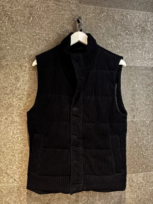 Marinblå manchester dunväst från dressman - Säljer en marinblå dunväst i manchester med hög krage och quiltad design. Västen har en clean look utan synliga knappar eller dragkedja framtill och är perfekt för lite kyligare dagar! 