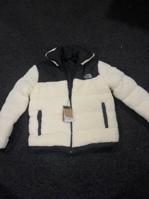 Vit teddyjacka från The North Face - Säljer en fet teddyjacka från The North Face i vit och svart. Jackan har hög krage, dragkedja framtill och två sidofickor med svarta detaljer. Materialet är mjuk fleece och syntet, perfekt för kalla dagar. Loggan är broderad på bröstet och jackan har en schysst puffer-look.