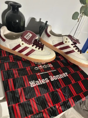 Adidas Wales Bonner Samba sneakers - Adidas Wales Bonner Samba sneakers i beige och vinrött med klassiska tre ränder på sidan. Skorna har en platt gummisula, snörning och detaljerad tunga med Wales Bonner-logga. Materialmix av skinn och textil, med kontraststickningar och retrovibe.