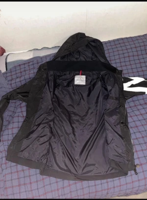 Svart vindjacka från Moncler med huva - Svart vindjacka från Moncler med huva och dragkedja framtill. Jackan har en stor vit zigzag-detalj på ena ärmen och Moncler-logga. Tillverkad i lätt polyester, perfekt för blåsiga dagar. Snygg och sportig look med långa ärmar och praktiska fickor.