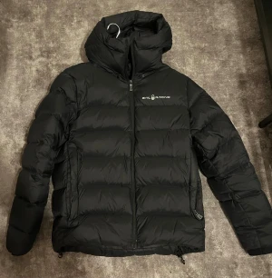 Svart pufferjacka från Sail Racing - Svart pufferjacka från Sail Racing med huva och diskret logga på bröstet och ryggen. Jackan har dragkedja framtill, quiltad design och två sidofickor. Perfekt för kalla dagar och har en clean, sportig look som funkar till streetwear. Jackan är i väldigt bra skick