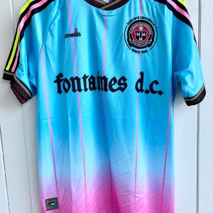 Fontaines d.c. - Fotbollströja - Merchandise - Säljer en Fontaines d.c. fotbollströja i ljusblå och rosa färgtoner. På framsidan finns texten ”fontaines d.c.” tryckt i svart, samt ett klubbmärke med texten ”DUBLIN'S ORIGINALS SINCE 1890”.   Baksidan av tröjan har texten ”i thought it was love” tryckt i limefärg. Nedtill på tröjan finns ett märke med en palestinsk flagga.  Tröjan är i nyskick och aldrig använd.  Storlek: XL