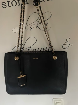 Svart axelväska från DKNY med kedja - Snygg svart axelväska från DKNY med guldfärgade kedjehandtag och detaljer. Väskan är tillverkad i skinn med en stilren, rektangulär form och har DKNY-logga i guld framtill. Perfekt storlek för att få plats med allt du behöver och har en elegant look.