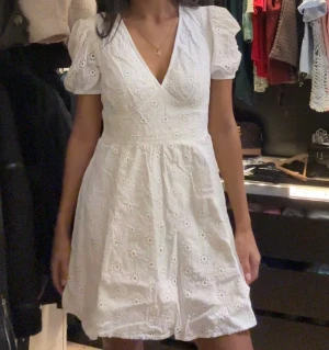 Vit kort klänning med puffärm, studentklänning  - Supersöt vit klänning med broderade blommor och puffiga korta ärmar. Klänningen har v-ringad hals och markerad midja som ger en fin siluett. Tillverkad i bomull, perfekt för varma dagar. Den har en kort längd och romantiska detaljer som gör den extra charmig.