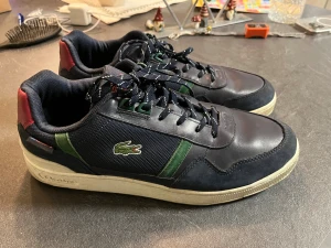 Mörkblå sneakers från Lacoste - Snygga mörkblå sneakers från Lacoste med gröna detaljer och röd häl. Klassisk krokodillogga på sidan och bak. Skorna har snörning, platt sula och rund tå. Materialmix av skinn och syntet med meshpartier för extra komfort. Perfekt för dig som gillar sportig stil.