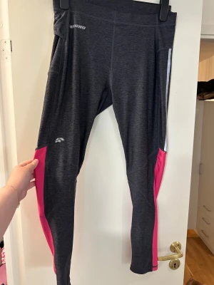 Träningsbyxor Karhu mörkgrå/rosa storlek: XL - Snygga träningsbyxor från Karhu i mörkgrått med rosa och vita detaljer längs benen. Stretchigt syntetmaterial som sitter tight och bekvämt. Dragkedja vid benslut och reflekterande ränder för extra synlighet. Perfekta för löpning eller gymmet.