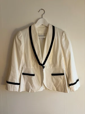 Vit kavaj med svarta detaljer Stockh LM - Snygg vit kavaj från Stockh LM i storlek 34 med svarta kontrastdetaljer längs krage, fickor och ärmslut. Kavajen har en knapp framtill, två fickor och lätt puffade axlar. Materialet är bomull och polyester, vilket ger en mjuk men stadig känsla.