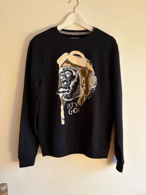 Svart sweatshirt med tryck från Blend - Svart sweatshirt från Blend i storlek M med coolt tryck framtill. Tröjan har rund hals, ribbade muddar och är långärmad. Materialet är mjuk bomull och polyester.