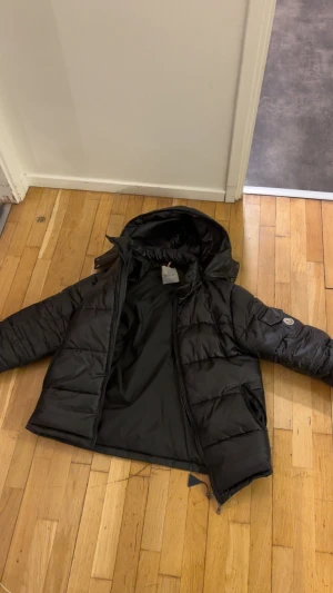 Svart Moncler pufferjacka med huva - Svart pufferjacka från Moncler med stor huva och klassisk logga på ärmen. Jackan har quiltad design, dragkedja framtill och praktiska fickor. Materialet är glansig polyester och dunfyllning som håller dig varm under kalla dagar. Perfekt för dig som gillar streetstyle.